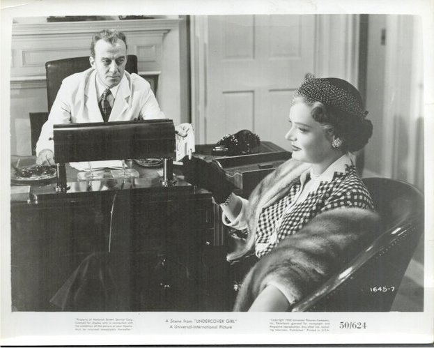 Black-and-white still of a man in a white coat at a desk, facing a woman in a checked coat and fur stole seated nearby, in a vintage setting.