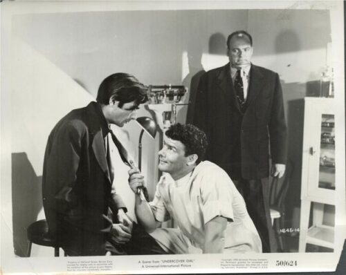 A man in a white medical uniform uses a stethoscope on the chest of a seated man in a dark suit, as another suited man stands in the background.