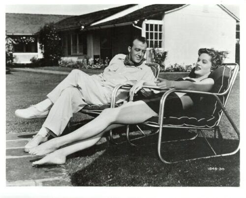 A man and woman in mid-century clothing relaxing on metal lawn chairs in a suburban yard, chatting laid-back in front of a house.