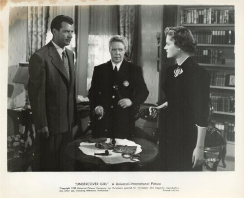 Three people in a 1940s interior: a man in a suit, a female police officer, and another woman in a dark dress stand around a table with papers and small objects.