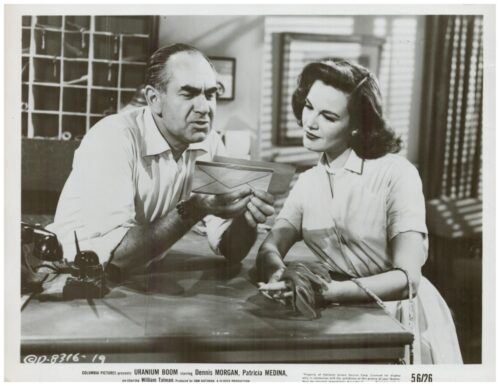 A man and a woman in 1950s attire lean over a desk, examining a letter together in a vintage office setting, black-and-white photo.