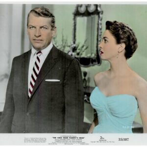 Mid-century movie still: a man in a dark suit and striped tie stands beside a woman in a turquoise strapless dress indoors, both looking in different directions.