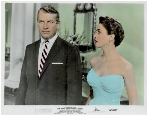 Mid-century movie still: a man in a dark suit and striped tie stands beside a woman in a turquoise strapless dress indoors, both looking in different directions.