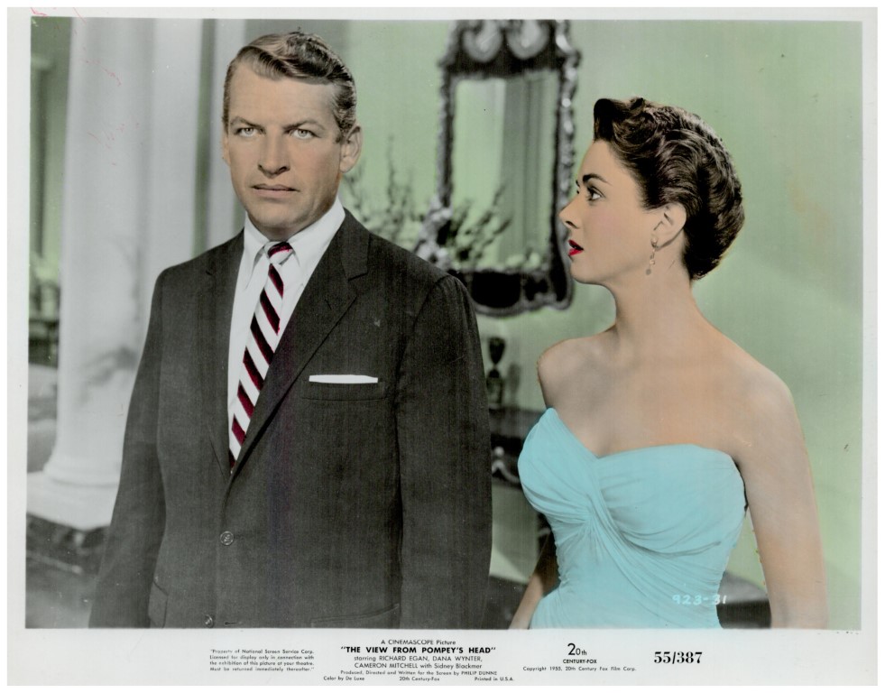 Mid-century movie still: a man in a dark suit and striped tie stands beside a woman in a turquoise strapless dress indoors, both looking in different directions.