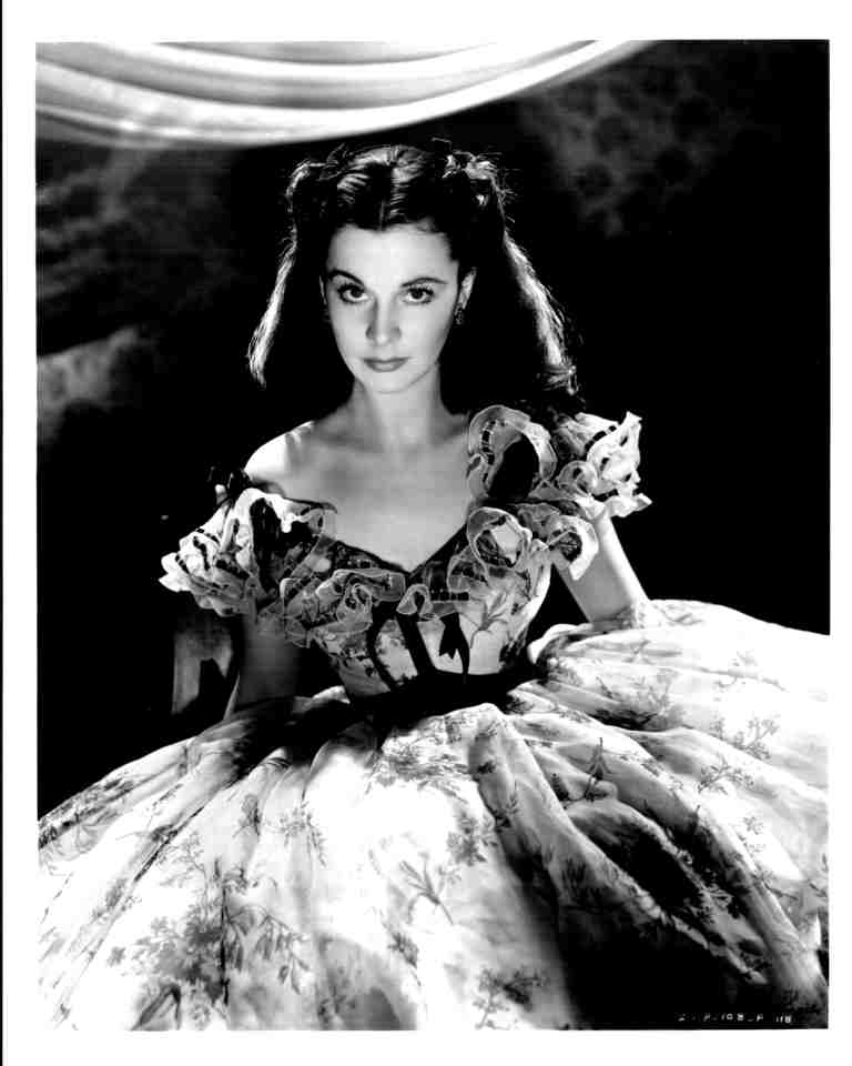 Black-and-white portrait of a woman in an elaborate ball gown with floral embroidery, looking directly at the camera.