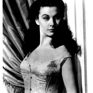 Black-and-white portrait of a woman with dark hair wearing a lace-trimmed corset, posing beside draped curtains.