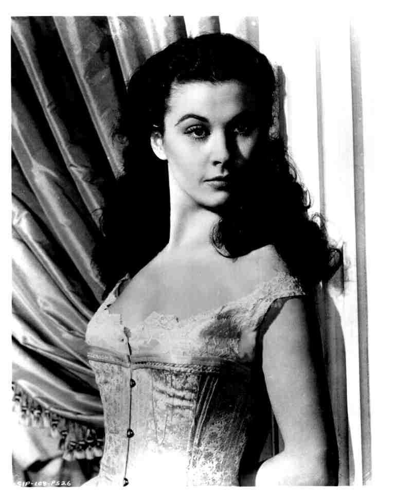 Black-and-white portrait of a woman with dark hair wearing a lace-trimmed corset, posing beside draped curtains.