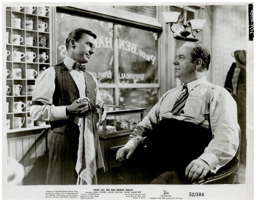 Production still from 'Wait Till the Sun Shines, Nellie': a young man in a vest and bow tie talks with a seated man in suspenders inside a shop.