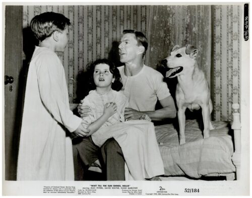 A man sits on a bed with a young girl on his lap as a boy stands nearby, while a dog stands beside them in a bedroom scene.