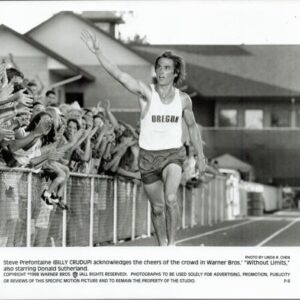 Steve Prefontaine in an Oregon running singlet raises his arm as fans cheer behind a track fence.