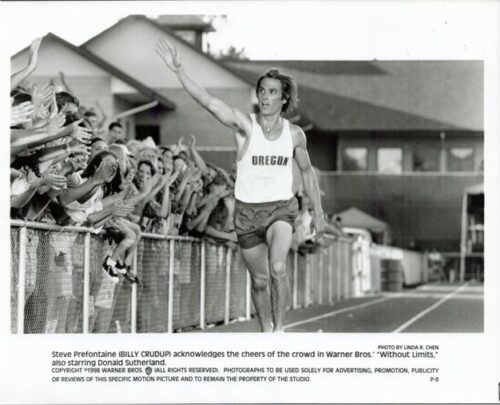 Steve Prefontaine in an Oregon running singlet raises his arm as fans cheer behind a track fence.