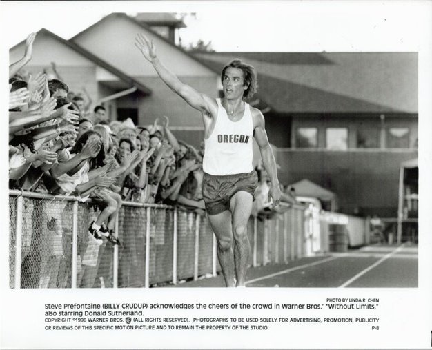 Steve Prefontaine in an Oregon running singlet raises his arm as fans cheer behind a track fence.