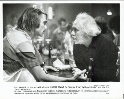 Two men lean toward each other over a table on a film-set bar, talking as crew and lights loom in the background.