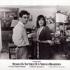 Two characters converse in a cluttered living room, with shelves and equipment in the background; a scene from Women on the Verge of a Nervous Breakdown.
