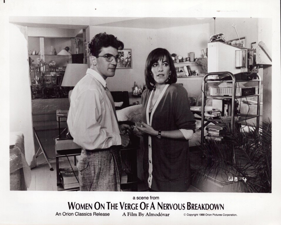 Two characters converse in a cluttered living room, with shelves and equipment in the background; a scene from Women on the Verge of a Nervous Breakdown.