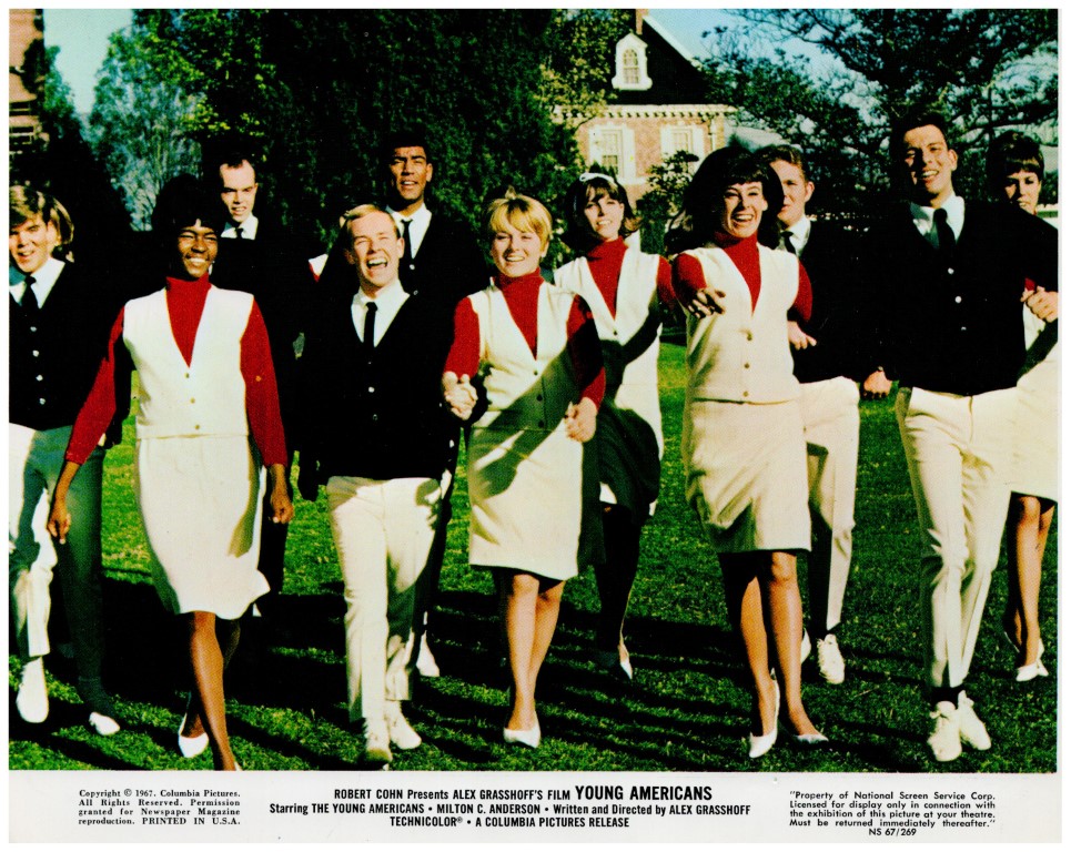Group of young adults in vintage uniforms walking toward the camera on a sunny green lawn, smiling together.