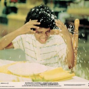 A young man in a striped shirt sits at a table, hands on his head as pie or whipped cream bursts onto him and the plate of food sits nearby.