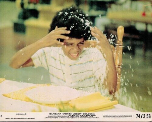 A young man in a striped shirt sits at a table, hands on his head as pie or whipped cream bursts onto him and the plate of food sits nearby.
