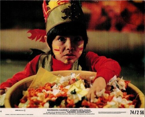 Child in a feathered headdress and red outfit with face paint, reaching into a large bowl of assorted candy.