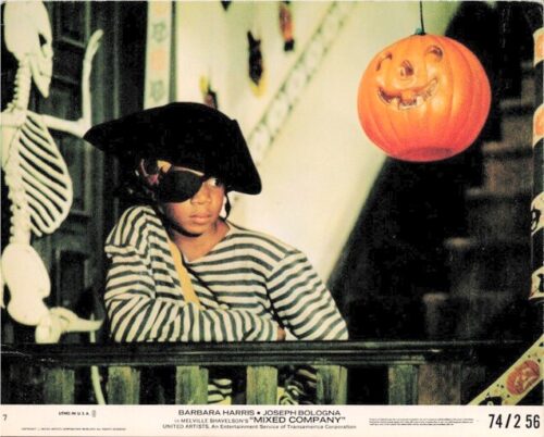 Child dressed as a pirate with striped shirt, eye patch and hat leans on a front porch railing during Halloween; a carved jack-o'-lantern hangs nearby.