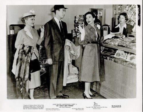 Black-and-white 1940s store scene: a man in a suit and hat stands with a coat over his arm, flanked by two women in period dresses, one at a counter speaking to a clerk.