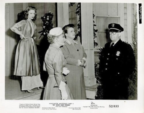 Two women in 1950s dresses converse with a uniformed police officer on a porch as a third woman watches in the background.