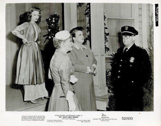 Two women in 1950s dresses converse with a uniformed police officer on a porch as a third woman watches in the background.