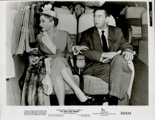 Two passengers sit in an airplane cabin: a stylish woman in a hat and fur stole beside a man in a suit, both looking to the left.