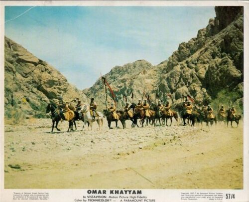 Line of riders on horseback crossing a rocky desert canyon, carrying banners, under a blue sky (cinema lobby card for Omar Khayyam).