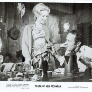 Black-and-white film still: a woman stands by a cluttered table as a man sits and looks up at her in a rustic room set.
