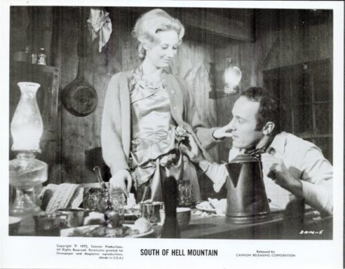 Black-and-white film still: a woman stands by a cluttered table as a man sits and looks up at her in a rustic room set.