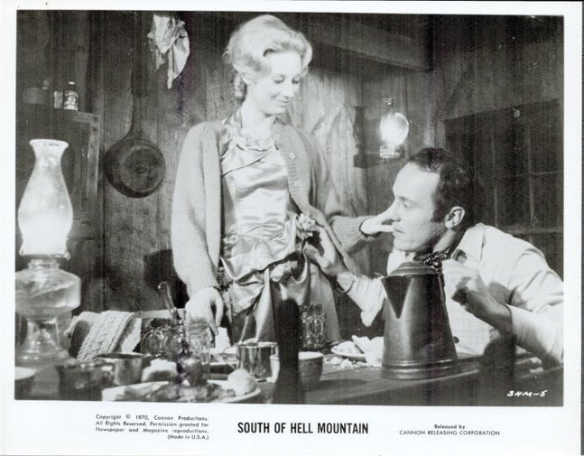 Black-and-white film still: a woman stands by a cluttered table as a man sits and looks up at her in a rustic room set.