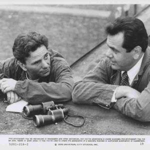 Two men in casual jackets lean on a concrete ledge, talking, with a pair of binoculars on the ground nearby.