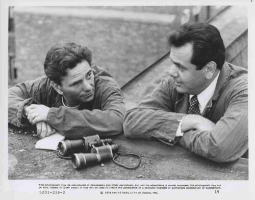 Two men in casual jackets lean on a concrete ledge, talking, with a pair of binoculars on the ground nearby.