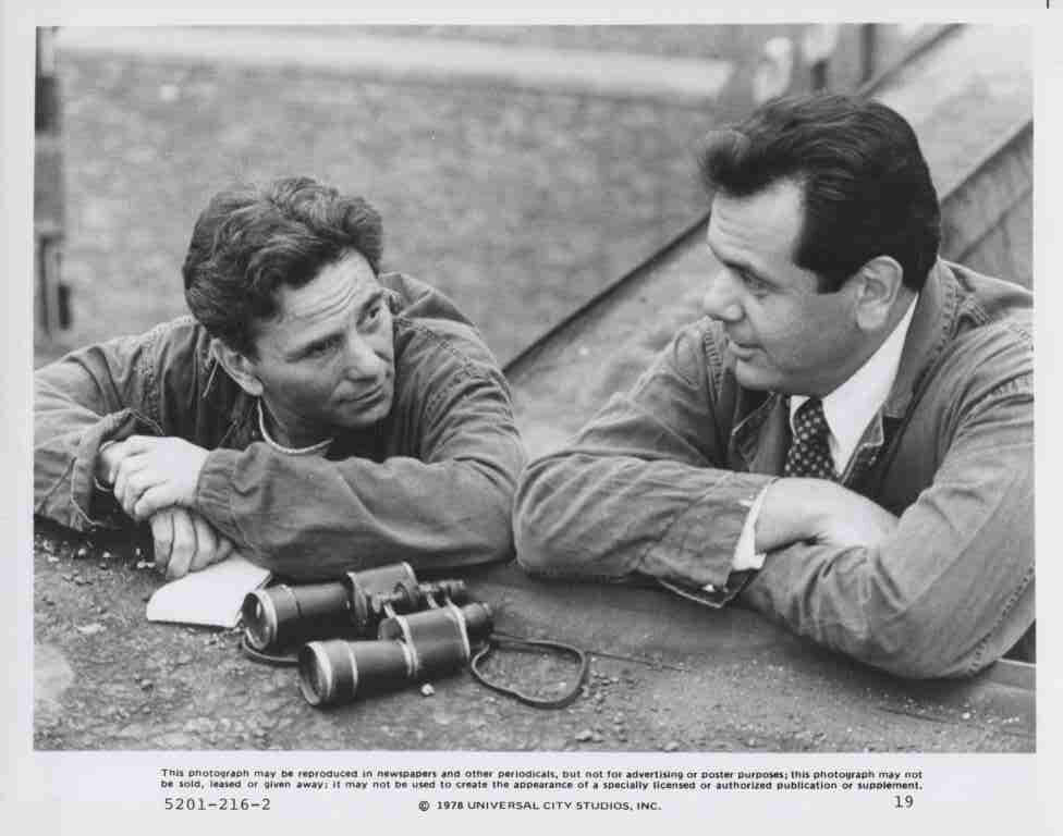Two men in casual jackets lean on a concrete ledge, talking, with a pair of binoculars on the ground nearby.