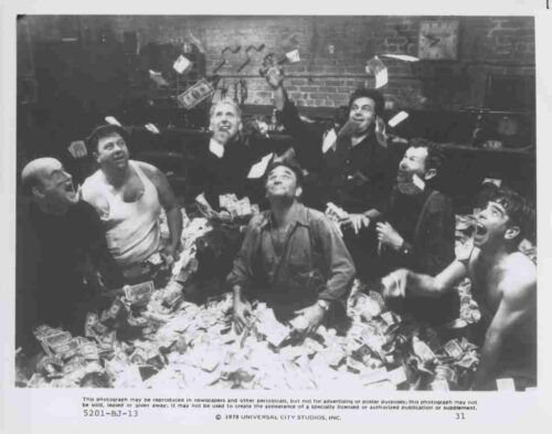 Eight men in a money-strewn room throw cash into the air, smiling as bills flutter around them.