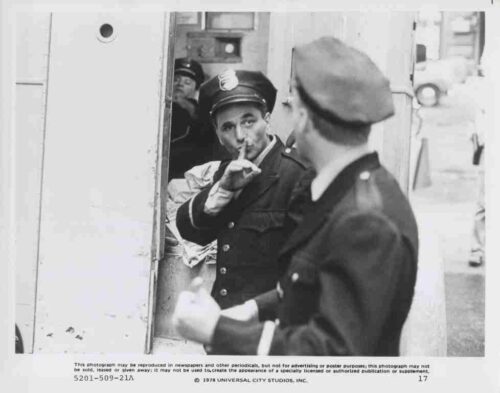 Policeman in uniform gesturing for silence with a finger to his lips, standing in a doorway while another officer faces him.