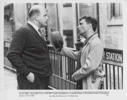 Two men chat on a city street near a subway station entrance; one man holds a hat toward the other as they talk in a casual pose.