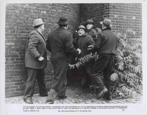 Group of men in coats and hats restrain a woman against a brick wall outdoors.