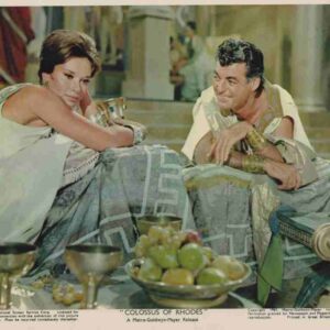 Movie still: two characters in ancient robes recline on cushions at a banquet, with fruit bowls and goblets in the foreground, from Colossus of Rhodes.