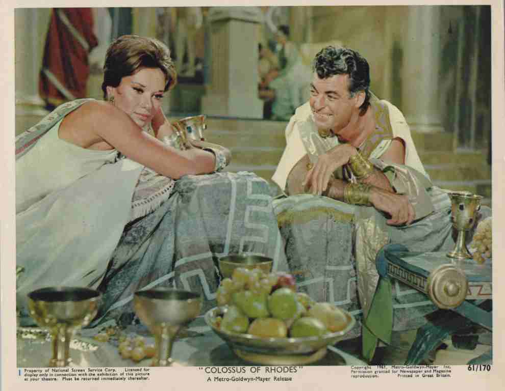 Movie still: two characters in ancient robes recline on cushions at a banquet, with fruit bowls and goblets in the foreground, from Colossus of Rhodes.