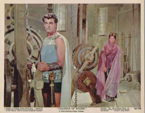 Still from the 1961 film Colossus of Rhodes: a muscular hero in armor and cape stands in a workshop, with a veiled woman in pink in the background.