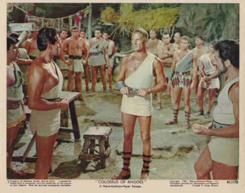 Colossus of Rhodes scene: muscular man in a white tunic with leather leg guards faces a crowd of warriors in ancient Greek attire.