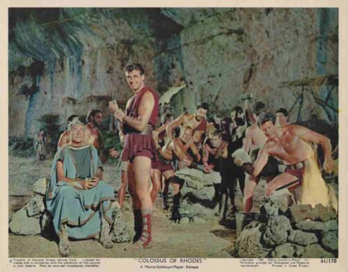 Film still: muscular warriors in ancient garb forge weapons in a rocky cave, central figure holding a dagger.