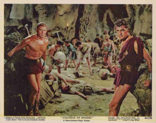 Two shirtless warriors in ancient armor stand at the edge of a rocky cavern as a chaotic battle unfolds behind them.