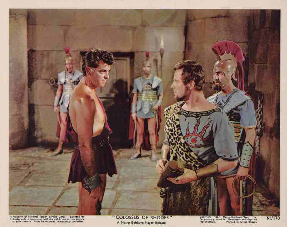 Two warriors face off in a stone arena as guards in armor observe from behind them at close range.