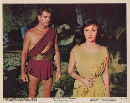 Film still: a man in a maroon one-shoulder tunic and belt stands beside a woman in a gold gown on a rocky outdoor set, both looking ahead.