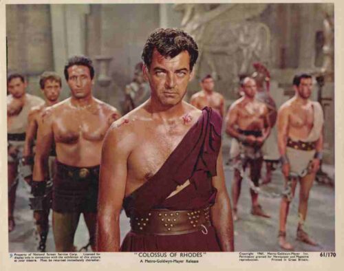 Muscular man in a gladiator outfit stands center, with others in loincloths behind him on a historic set — a film still from Colossus of Rhodes (1954).