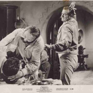 Two men wrestle on a table in a black-and-white studio scene while a third man watches, a prop sword on his head,”