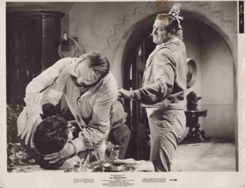 Two men wrestle on a table in a black-and-white studio scene while a third man watches, a prop sword on his head,”